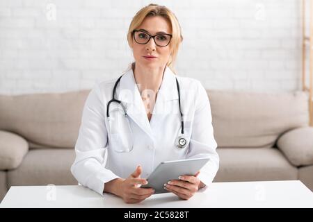 Bella donna adulta che lavora online da casa. Medico con bicchieri e cappotto bianco tiene la compressa in mani Foto Stock