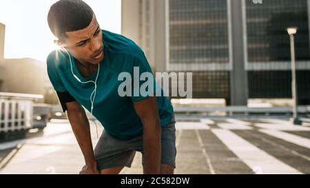 Atleta maschile che si appoggia in avanti con le mani sulle ginocchia e guarda lontano in città. Uomo corridore che fa una pausa dopo la corsa di mattina. Foto Stock