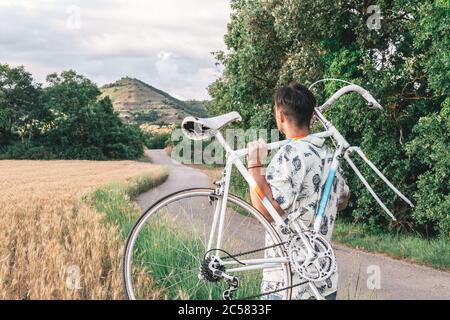 Bel giovane uomo porta una moto senza ruota. Indossa una camicia alla moda. È nel mezzo di una strada forestale per un polecat. Foto Stock