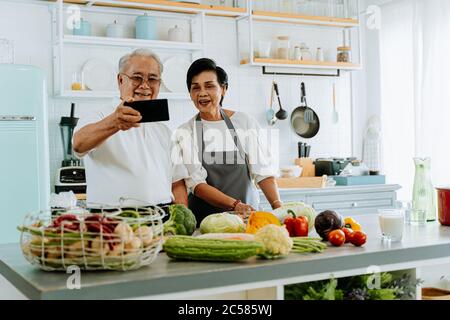 Allegro anziano uomo e donna asiatici sorridendo e salutando amico mentre si effettua una videochiamata su smartphone e cucinare in cucina Foto Stock