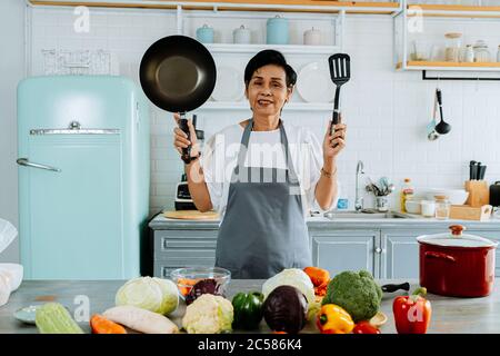 Felice femmina asiatica senior in grembiule sorridente per la macchina fotografica e mostrando padella e spatola mentre si prepara il pranzo in cucina a casa Foto Stock