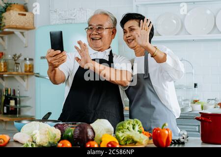 Allegro anziano uomo e donna asiatici sorridendo e salutando amico mentre si effettua una videochiamata su smartphone e cucinare in cucina Foto Stock