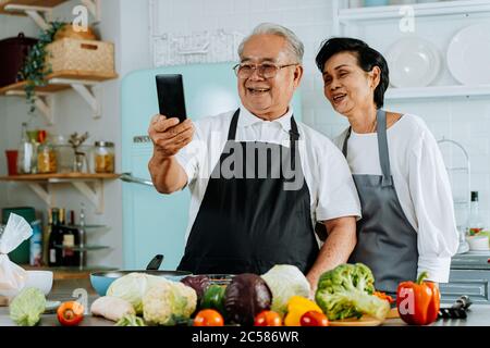 Allegro anziano uomo e donna asiatici sorridendo e salutando amico mentre si effettua una videochiamata su smartphone e cucinare in cucina Foto Stock
