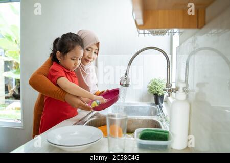 Aiuto mano. Cute Little Girl aiuta la sua madre musulmana nel lavare i piatti alla cucina di famiglia Foto Stock