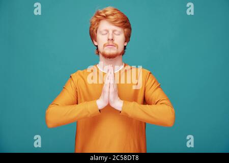 Calma spirituale giovane uomo rosso con barba facendo gesto Namaste meditando con occhi chiusi su sfondo blu Foto Stock