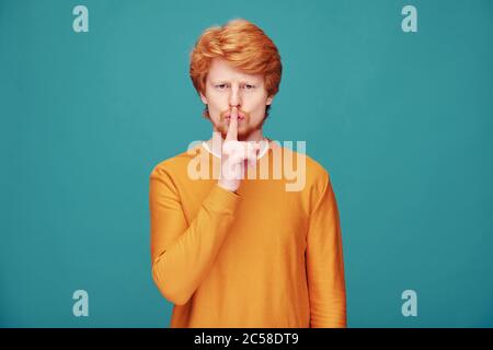 Bell'uomo rosso in maglia arancione che tiene il dito vicino alla bocca mentre chiede di mantenere la calma Foto Stock