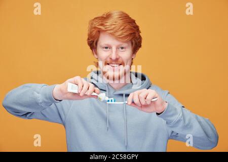 Ritratto di un bel giovane uomo rosso sorridente con felpa con cappuccio applicando dentifricio sullo spazzolino su sfondo arancione Foto Stock