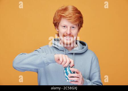 Ritratto di giovane allegro rosso uomo in felpa con cappuccio apertura bottiglia vitamina mentre si prende cura della sua immunità Foto Stock