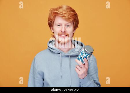 Ritratto di sorridente giovane rosso uomo in felpa con cappuccio prendendo vitamine per essere sano, sfondo arancione Foto Stock