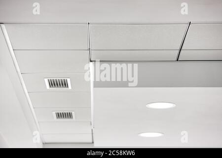 soffitto in cartongesso multilivello e soffitto sospeso in piastrelle bianche quadrate con lampade di illuminazione integrate e griglie di ventilazione. Foto Stock
