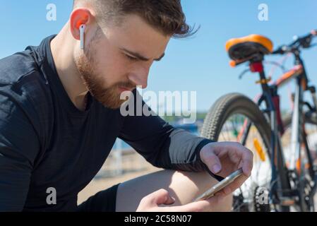 Ritratto di un atleta di ciclista che riposa dopo un giro in bicicletta usando uno smartphone in mano. All'aperto. Sport primaverili. Il ragazzo ha venticinque anni Foto Stock