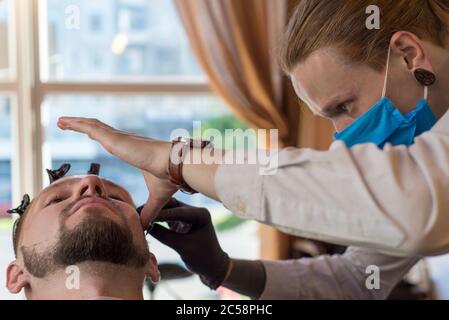 un parrucchiere taglia un giovane ragazzo con un rifinitore per capelli, pettinando i capelli sulla sua testa. Lavoro del maestro nel taglio di capelli degli uomini in un barbiere Foto Stock