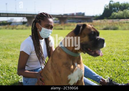 Fuoco selettivo su una donna africana etnica che indossa una maschera protettiva e seduto accanto ad un cane di boxer sull'erba con un ponte sullo sfondo Foto Stock