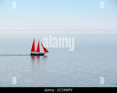 Una barca con tre vele rosse, galleggianti su un mare limpido Foto Stock