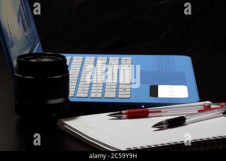 Chiusura laterale di un computer portatile blu aperto, obiettivo della fotocamera e due penne rosse e nere su un notebook vuoto Foto Stock