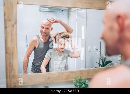 Papà calvo e il suo figlio adolescente dai capelli lunghi in bagno di fronte allo specchio. Padre mostrando a ragazzo il suo nuovo taglio di capelli stile. Stile di vita familiare comune e. Foto Stock