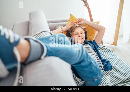 Sorridente ragazzo di Preteen che si trova nel salotto domestico pieno di luce del sole su divano accogliente vestiti jeans casual e sneakers ascoltare la musica utilizzando wireless lui Foto Stock