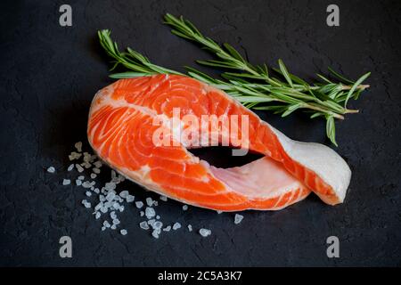 Bistecca di salmone crudo con rosmarino e sale marino su ghiaccio su fondo nero. Vista dall'alto. Concetto di cibo sano e dietetico. Dieta del keto. Pesce rosso grezzo preparato per Foto Stock