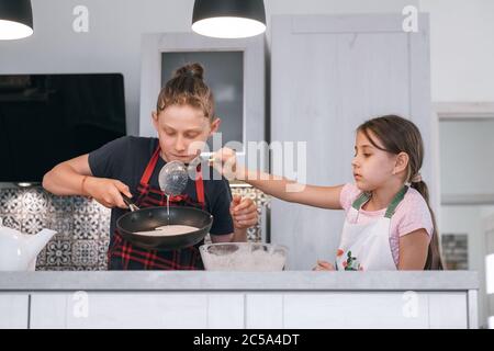 Fratello e sorella hanno vestito grembiuli facendo una frittella fatta in casa sulla cucina. Ragazza porendo un impasto liquido sulla padella calda. Concetto di cucina casalinga per bambini Foto Stock