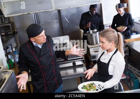 Disorientati chef rendendo impotente gesto mentre parla con sorpresa la cameriera che puntano a piatto cucinato Foto Stock