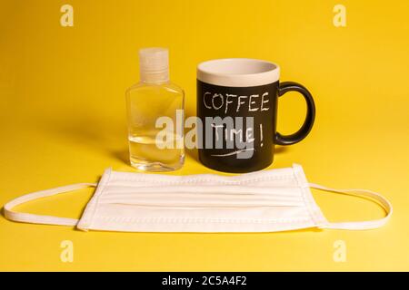Tazza di caffè con parole scritte in gesso accanto ad una bottiglia di gel disinfettante e una maschera protettiva contro le malattie su sfondo giallo. Concetti Foto Stock