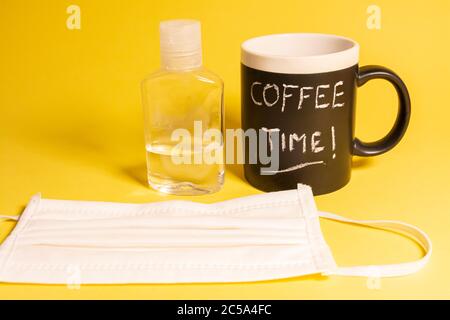 Tazza di caffè con parole scritte in gesso accanto ad una bottiglia di gel disinfettante e una maschera protettiva contro le malattie su sfondo giallo. Concetti Foto Stock