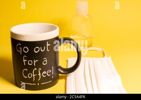 Tazza di caffè con parole scritte in gesso accanto ad una bottiglia di gel disinfettante e una maschera protettiva contro le malattie su sfondo giallo. Concetti Foto Stock