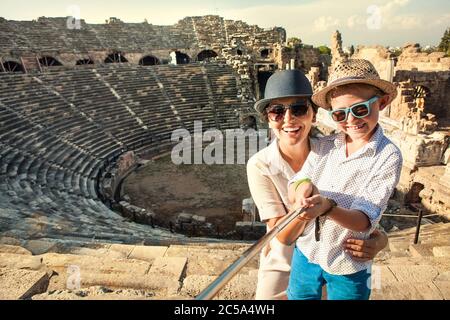 Madre con figlio sorridente alla fotocamera scattando una foto selfie in un teatro antico utilizzando un bastone selfie. Viaggiare in tutto il mondo con l'immagine concettuale dei bambini. Foto Stock