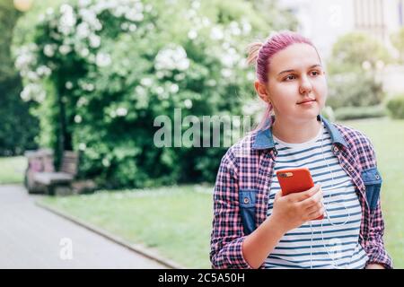 Ritratto di bella giovane giovane ragazza sorridente moderna in una camicia a scacchi ascoltando musica utilizzando smartphone e auricolari mentre cammina nel parco. Foto Stock