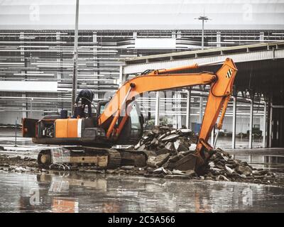 Il trattore Caterpillar rastrema grandi pezzi di cemento durante il tempo nuvoloso sullo sfondo di un grande edificio Foto Stock