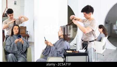 Positivo per il parrucchiere maschile che fa acconciatura per la donna giovane, donna che usa il telefono cellulare Foto Stock