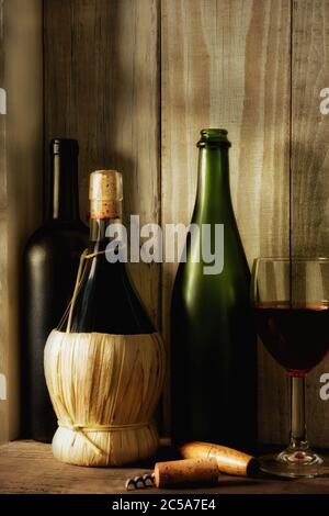 Un vino ancora vivo con luce calda da una finestra sul lato. Tre bottiglie, un bicchiere di vino e una vite di sughero in un ambiente rustico, formato verticale, Foto Stock