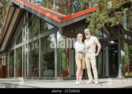 Bella coppia senior in piedi contro cottage di vetro e abbracciarsi l'un l'altro all'aperto Foto Stock