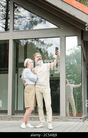 Bell'uomo anziano che abbracciava la moglie mentre prendeva selfie con lui vicino a casa di vetro Foto Stock