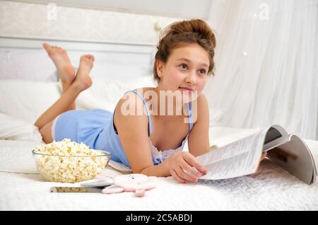 La ragazza è sdraiata sul letto in pigiama guarda una rivista. Adolescenti di passatempo. Tempo libero fuori dalla scuola. Durata della quarantena. Foto Stock