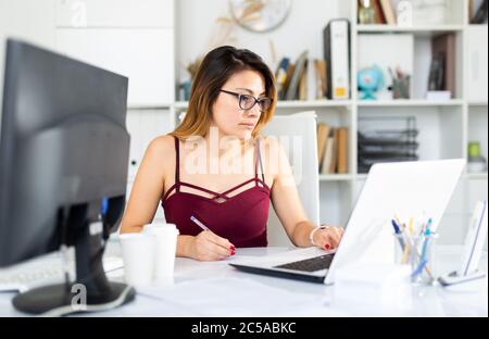 Donna latina positiva in abbigliamento casual che lavora da sola con laptop e carte in ufficio Foto Stock