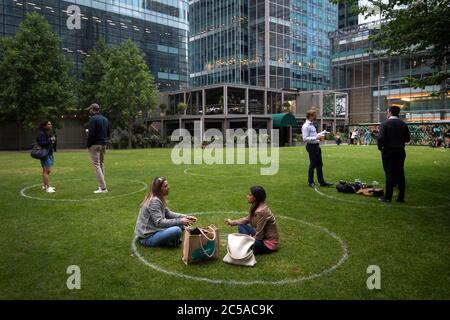 I bevitori si trovano all'interno di anelli segnati sull'erba per mantenere la loro distanza sociale fuori da un bar a Canary Wharf, Londra Est, mentre ulteriori restrizioni di blocco del coronavirus sono revocati in Inghilterra. Foto Stock