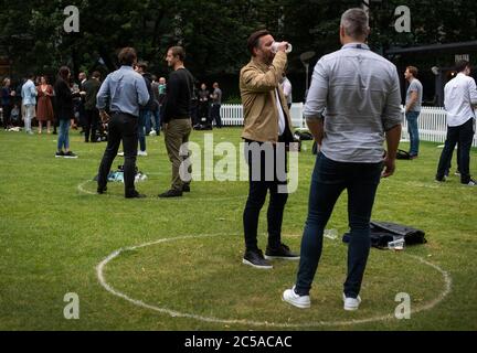 I bevitori si trovano all'interno di anelli segnati sull'erba per mantenere la loro distanza sociale fuori da un bar a Canary Wharf, Londra Est, mentre ulteriori restrizioni di blocco del coronavirus sono revocati in Inghilterra. Foto Stock