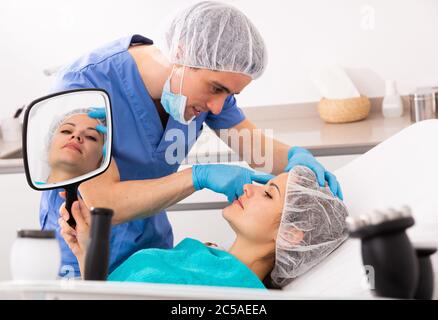 Giovane cliente femminile che guarda lo specchio mentre estetista qualificato che esamina la sua pelle del viso dopo la procedura in studio di cosmetologia medica Foto Stock