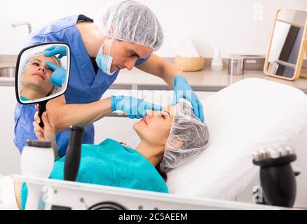 Giovane cliente femminile che guarda lo specchio mentre estetista qualificato che esamina la sua pelle del viso dopo la procedura in studio di cosmetologia medica Foto Stock