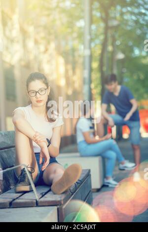 Ritratto di ragazza adolescente carina in occhiali seduti su panchina sulla strada della città, godendo di calda giornata estiva Foto Stock