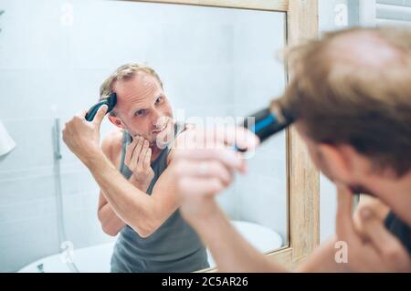Concetto di trattamento corpo e pelle. Smiling Man creare un taglio di capelli nuovo stile rifinire un pelo usando un rifinitore elettrico ricaricabile che guarda in bagno Foto Stock