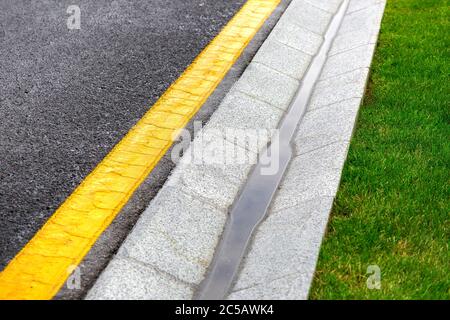 sistema di drenaggio bordo con acqua piovana in prossimità di una strada asfaltata con una marcatura gialla e un prato verde dopo la pioggia. Foto Stock