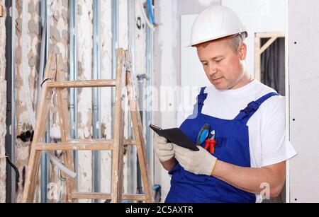 Allegro imprenditore maschile positivo che ha rottura in interni cantiere, appoggiandosi a parete con telefono in mani Foto Stock