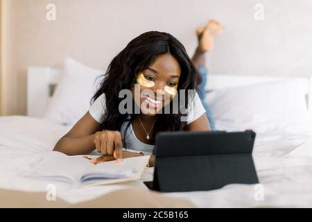 Sorridente donna africana si trova sul letto, scorrendo attraverso il suo computer tablet, lavorando online, facendo compiti, preparando test o rilassandosi. Operazione riuscita Foto Stock