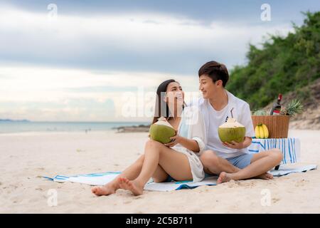 Giovane coppia asiatica seduta sulla coperta da picnic e bevendo acqua di cocco in spiaggia e vicino al mare con frutta tropicale in background. Estate, vacanze Foto Stock