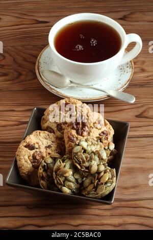 Biscotti di farinata d'avena con noci e uvetta, biscotti di semi di zucca ricoperti di zucchero in una ciotola quadrata e una tazza di tè nero su un tavolo di legno. Primo piano Foto Stock