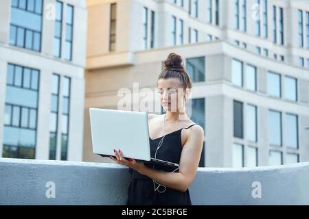bella ragazza cammina per la città, lavorando su un computer portatile, tenendo un computer portatile e ascoltando musica. Foto Stock