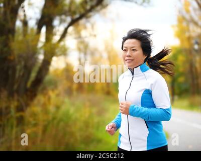 Donna attiva nel suo cinquanta corsa e jogging. Medio invecchiamento asiatico mature femmina jogger all'aperto vivere sano stile di vita in bellissimo parco della città d'autunno in colorato fogliame autunno. Cinese asiatico adulto nei suoi anni cinquanta. Foto Stock
