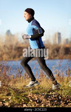 L'uomo che corre jogging in autunno ascoltando musica su smartphone. Allenamento dei corridori in outfit caldo nelle giornate fredde. Fit maschile fitness atleta modello di allenamento all'aperto in autunno. Lunghezza completa del corpo del pareggiatore. Foto Stock